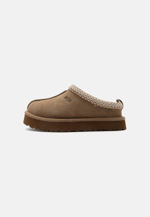Brun ruskind slip-on tøfler med tykk gummisål, strikket kant omkring åbningen og broderet mærkelogo på siden.