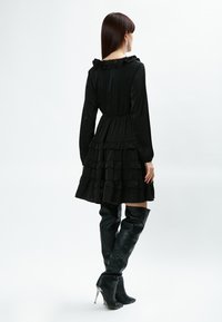 Vestido negro de manga larga con escote con volantes y falda en capas. Combinado con botas de cuero negro hasta la rodilla con punta afilada y tacones de aguja.