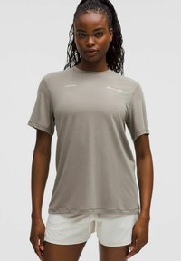 Frau mit geflochtenem Haar, die ein hellgraues T-Shirt mit kurzen Ärmeln und weiße Shorts trägt und vor einem einfarbigen Hintergrund steht.