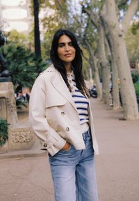 Žena s dlhými tmavými vlasmi, oblečená v krémovej bunde a pruhovanom svetri, stojí na chodníku lemovanom stromami v meste s rukami vreckách.