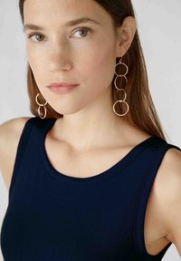 Gold circular drop earrings with three linked rings; model wearing a navy ribbed tank top, smooth texture, natural makeup, neutral background.