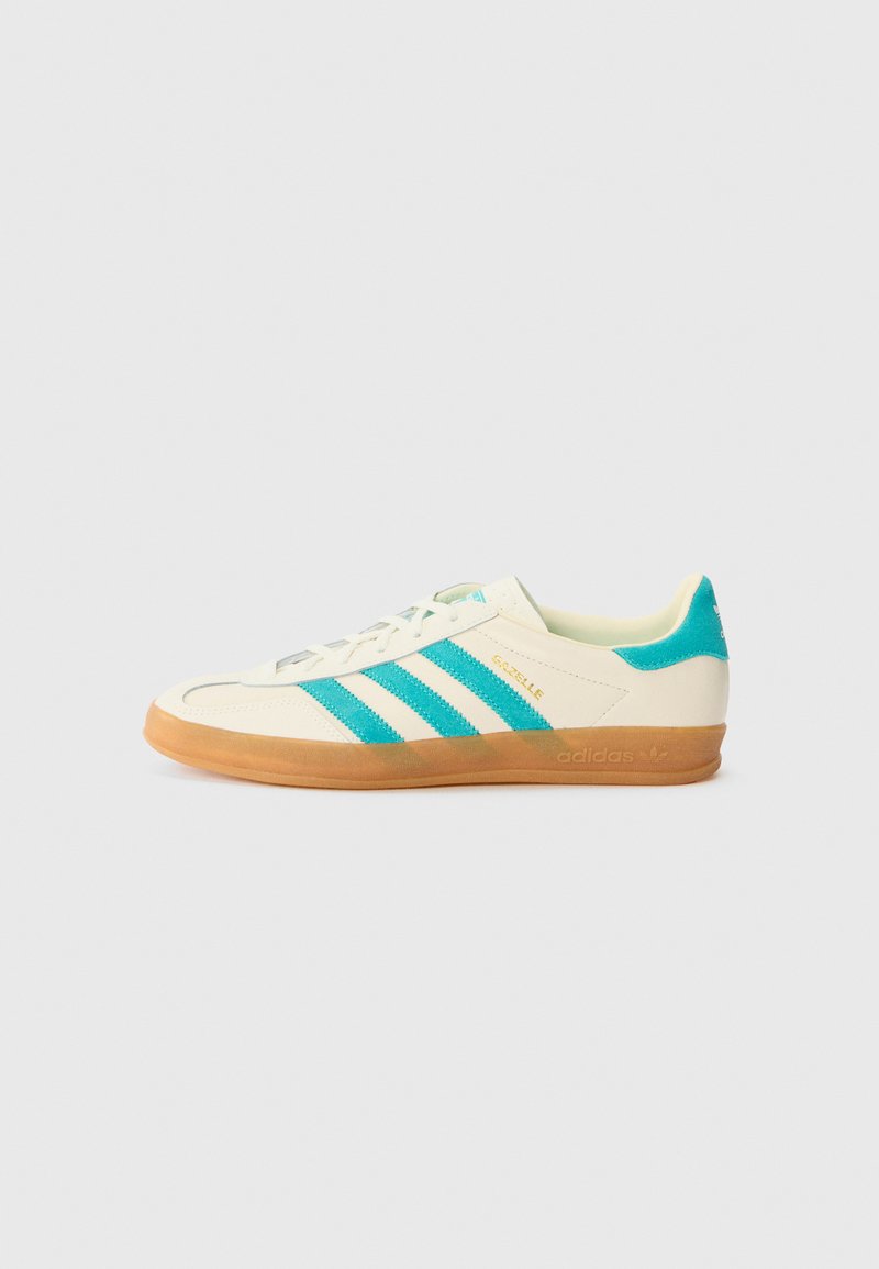 Witte sneaker met drie turquoise strepen, een gumzool en textuuraccenten. Kenmerkt zich door een lage profiel en een vetersluiting, gelabeld "Gazelle."