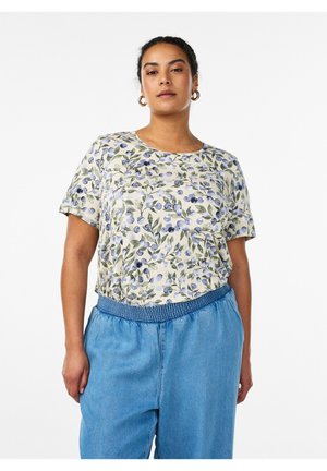 Woman wearing a short-sleeve floral top with purple berries and green leaves, paired with high-waisted blue denim pants.