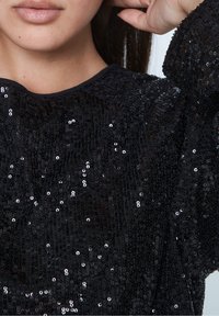 Top nero con paillettes e finitura testurizzata, con scollatura tonda e maniche morbide. Le paillettes creano un'effetto scintillante.