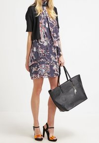 Svart axelväska med präglad blommönster, matchad med en mönstrad klänning i mörka nyanser och en kort svart cardigan. Klacksandaler med orange detaljer.