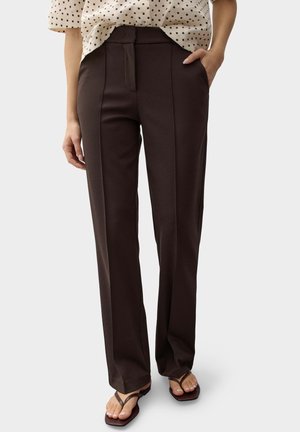 Donna che indossa pantaloni marrone scuro a gamba dritta con pieghe frontali, abbinati a una camicetta color crema con pois neri e sandali marroni.