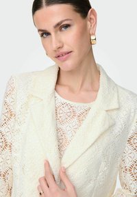 Bílý krajkový blejzr s květinovými vzory, texturovaná látka a klopený límec. Modelka má pod ním na sobě odpovídající krajkový top a náušnice ve tvaru kruhu.