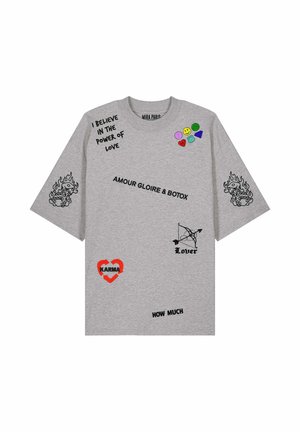Giovane uomo che indossa una maglietta grigia oversize con faccine sorridenti colorate, frasi nere, dadi infuocati, un cuore rosso con la scritta "KARMA" e un fiocco etichettato "Lover".