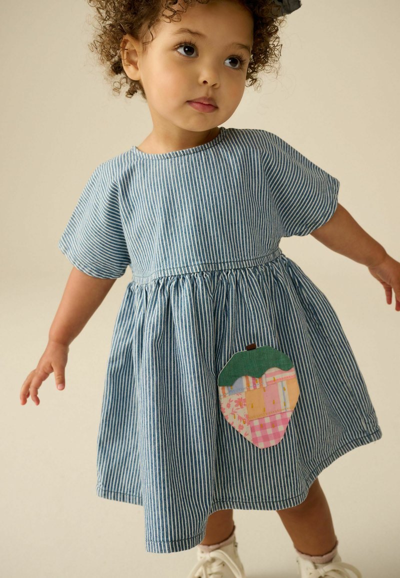 Bambino piccolo che indossa un vestito a righe blu e bianco con motivo fragola patchwork, in piedi con le braccia leggermente aperte su sfondo beige.