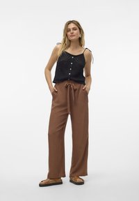 Top de punto negro con botones delanteros y tirantes ajustables, combinado con pantalones marrones de cintura alta con un lazo en la cintura y piernas anchas.