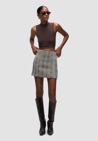 Bruine mouwloze top, gestructureerde zwart-witte houndstooth minirok en hoge bruine laarzen met een glanzende afwerking. Zonnebril en sieradenaccenten.