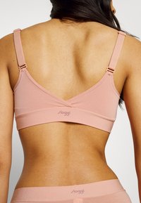 Light pink wireless bralette with adjustable straps, textured fabric, and a wide underband featuring a subtle logo at the back.