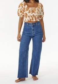 Femme portant un jean large taille haute bleu et un top court blanc avec un motif floral orange et des manches bouffantes, debout pieds nus.