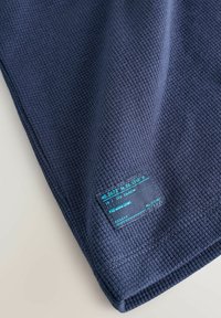 Tessuto blu navy con un motivo a nido d'ape testurizzato. Presenta un'etichetta rettangolare con coordinate e testo in turchese. Il materiale sembra leggero.