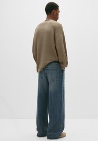 Jonge man gezien van achteren, gekleed in een losse beige trui, wijde blauwe jeans en beige schoenen, staand tegen een witte achtergrond.