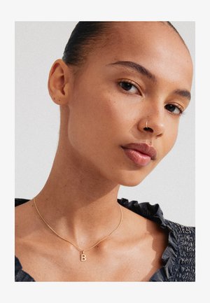 Guldkæde med et vedhæng formet som bogstavet "B," båret af en person mod en lys baggrund; struktureret mørk tøj med bølgede kanter.