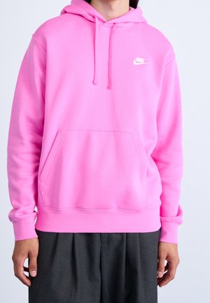 Person iført en skrigende pink Nike hættetrøje med frontlomme og mørkegrå bukser, stående mod en ensfarvet lys baggrund.