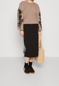 Pull marron avec broderie florale noire sur les manches, associé à une jupe midi noire et des baskets noires. Tenant un sac fourre-tout beige.