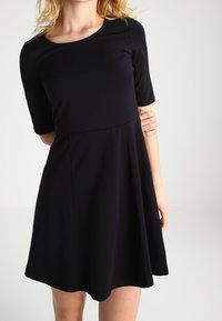 Kvinna klädd i en enkel svart kortärmad A-linje klänning, står mot en ljusgrå bakgrund.