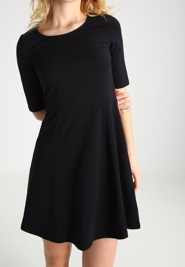 Kvinna klädd i en enkel svart kortärmad A-linje klänning, står mot en ljusgrå bakgrund.