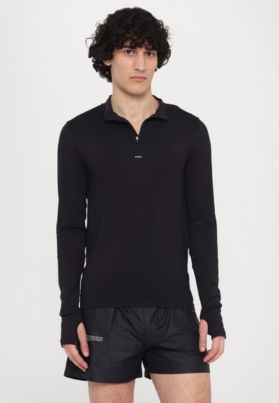 Schwarzes Langarmshirt mit halbem Reißverschlusskragen, ausgestattet mit Daumenlöchern an den Ärmeln. Kombiniert mit schwarzen Sportshorts, die ein minimalistisches Design aufweisen.