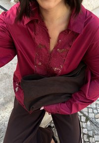 Rotbluse mit floralen Spitzenakzenten am Kragen und an den Ärmeln, kombiniert mit einer braunen Stoff-Clutch. Beiger Stein-Hintergrund.