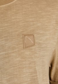 Tissu tricot beige avec un petit patch rectangulaire cousu présentant un design minimaliste à lignes blanches sur la zone de la poitrine.