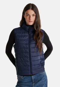 Žena s dlouhými hnědými vlasy má na sobě prošívanou námořnickou vestu přes černé tričko s dlouhým rukávem a modré džíny, stojí s rukama za zády.