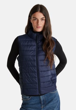Femme aux longs cheveux bruns portant un gilet matelassé bleu marine sur un t-shirt noir à manches longues et un jean bleu, se tenant avec les mains derrière le dos.