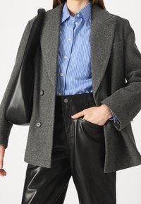 Šedé sako z vlněné směsi s volnějším střihem, dvouřadé zapínání a klopami s výřezem, doplněné modrou proužkovanou košilí a černými koženými kalhotami.