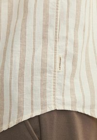 Camisa a rayas con rayas beige y marrón claro, confeccionada en tela ligera. Cuenta con una etiqueta que dice "ORIGINALS" cerca del dobladillo.