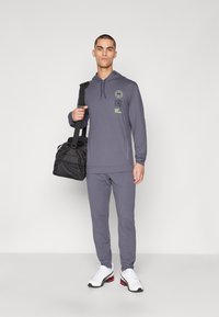 Conjunto deportivo para hombre en gris oscuro, que incluye una sudadera con capucha y detalles del logo, pantalones a juego y zapatillas blancas. El modelo lleva un bolso negro.