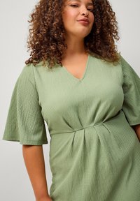 Grünes, strukturiertes Kleid mit kurzen, ausgestellten Ärmeln, V-Ausschnitt und Taillenraffung, gefertigt aus leichtem Material.