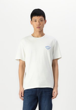 T-shirt a maniche corte di un leggero colore beige chiaro con scollo rotondo; presenta un logo blu sul lato sinistro del petto; abbinato a jeans blu scuro.