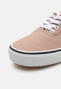 Baskets en toile beige vue de face avec lacets blancs, semelle en caoutchouc blanche et bande noire le long du bord de la semelle.