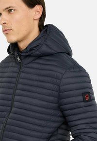 Doudoune bleu marine avec fermeture éclair à l'avant, design matelassé, capuche et accent en patch noir sur la manche gauche. Tissu lisse et léger.