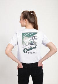 Bílý bavlněný tričko s vintage grafick�ým designem a zelenými akcenty, textem o rekordech v rychlosti a grafikou závodního auta.
