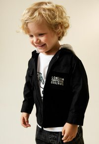 Črno jakno s sivim kapuco krasi žep na sprednji strani z napisom "LONDON FREESTYLE RIDER COOL VIBES", nošena je preko bele majice z grafiko in temnih kavbojk.