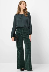 Mørkegrøn satinbluse med rund halsudskæring, kombineret med mørkegrønne teksturerede bredbenede bukser i ribstof med diamantmønster. Sort håndtaske.