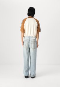 T-shirt raglan marron et crème avec deux rayures bleues, associé à un jean large bleu clair. Tissu texturé, modèle de dos.