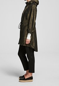 Giacca leggera da pioggia verde oliva, con vita elastica, cappuccio e coulisse; abbinata a pantaloni neri a sigaretta e zeppe espadrillas.