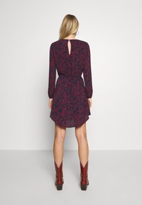 Robe bleu marine à motif floral rouge, manches longues, taille cintrée et détail dos en forme de goutte d'eau. Portée avec des bottes marron style cowboy.