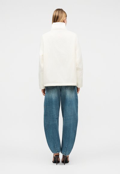 Γυναίκα που φοράει λευκό μπουφάν puffer, φαρδιά μπλε τζιν και μαύρα ψηλοτάκουνα σανδάλια, στέκεται πλάτη σε απλό φόντο.