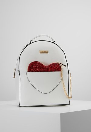 Biały plecak z czarnym wykończeniem, z przednią kieszenią w kształcie serca, w której znajduje się czerwony, błyszczący, sercowaty woreczek z złotym suwakiem na łańcuszku.