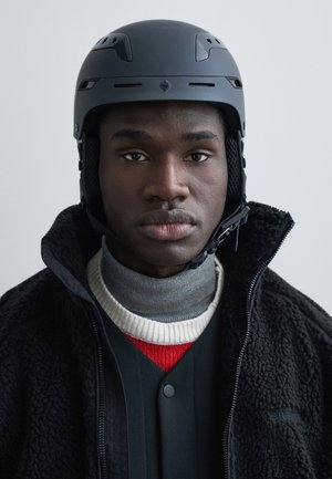 Νεαρός άνδρας που φοράει μαύρο κράνος ασφαλείας, μαύρο φλις μπουφάν πάνω από γκρι ζιβάγκο και κόκκινο-άσπρο πλεκτό πουλόβερ, κοιτάζει μπροστά με ουδέτερη έκφραση.