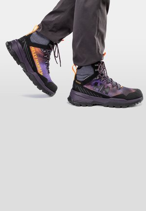 Botas de senderismo moradas y negras con tejido texturizado, suela de goma negra, acentos naranjas y un prominente logo de Helly Hansen en el lateral.