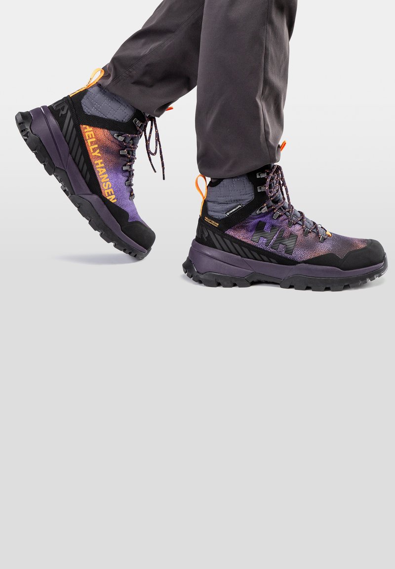 Scarpe da trekking viola e nere con tessuto testurizzato, suola in gomma nera, dettagli arancioni e logo Helly Hansen ben visibile sul lato.