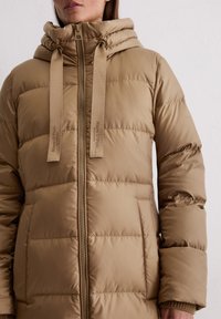 Bežs pūkains mētelis ar augstu apkakli, ar regulējamu kapuci, atklātu priekšējo rāvējslēdzēju un šūtu paneļu konstrukciju. Discreta zīmola emblēma uz kapuces auklām.