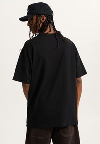 Schwarzes T-Shirt mit kurzen Ärmeln aus Baumwolle, mit lockerer Passform. Trägt eine passende schwarze Mütze. Einfaches Design ohne sichtbare Logos.