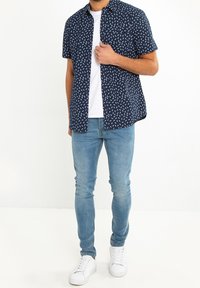 Korte mouwen marineblauwe overhemd met een wit bladerenpatroon, gecombineerd met lichtblauwe slim-fit jeans en witte sneakers.